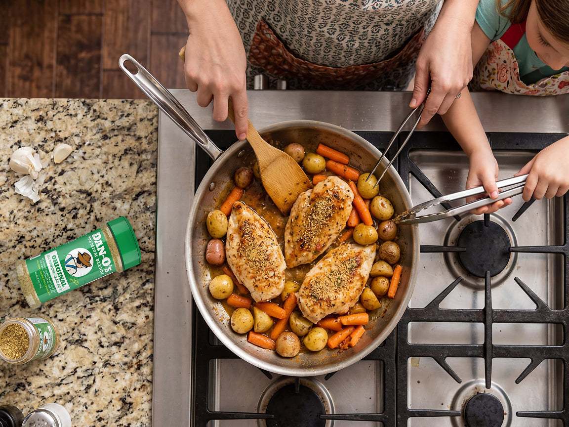 Overhead view of a stainless steel skillet on a gas stovetop, containing three pieces of seasoned chicken breast with roasted carrots and small potatoes. Two people in patterned aprons are visible from the shoulders down, using a wooden spoon and metal tongs to serve the food.